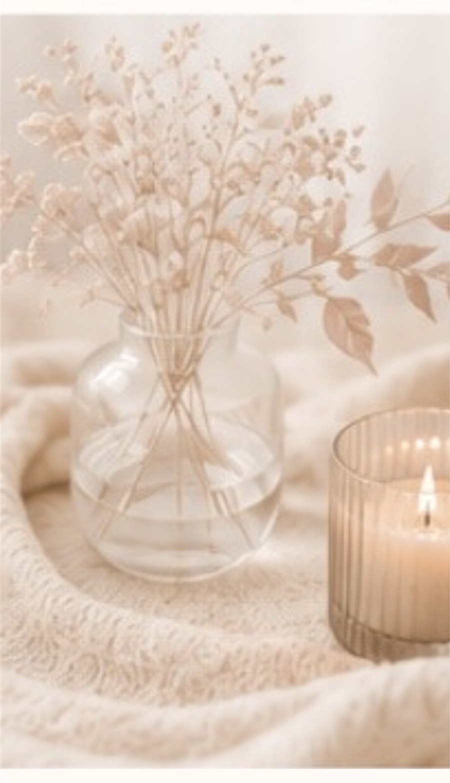 Glasvase mit hellen Trockenblumen und eine Kerze in warmem Licht.